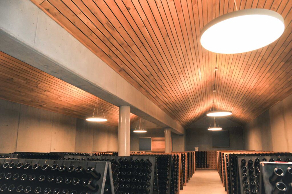 Ein Weinkeller mit hölzernen Decken, hellen Oberlichtern und Regalen voller Flaschen, die nach unten gekippt und in Reihen entlang der Betonwände angeordnet sind.