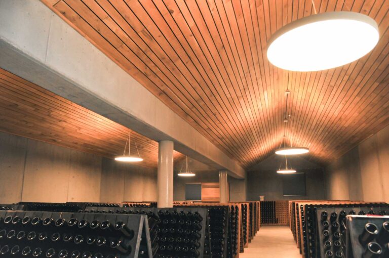 Ein Weinkeller mit hölzernen Decken, hellen Oberlichtern und Regalen voller Flaschen, die nach unten gekippt und in Reihen entlang der Betonwände angeordnet sind.