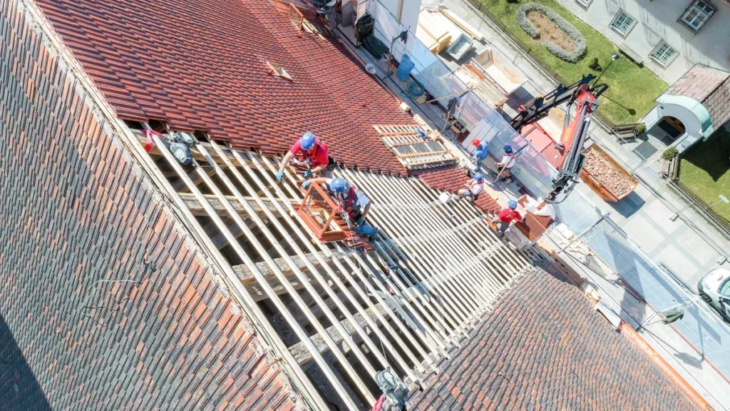 Eine Gruppe von Bauarbeitern mit Helmen repariert das Dach eines Gebäudes und ersetzt Holzbalken und Ziegel. Unten sind Maschinen und Geräte zu sehen, und die Szene wird von oben betrachtet.
