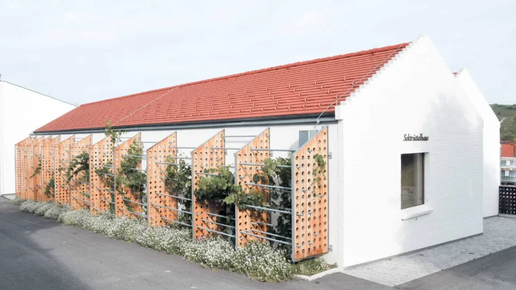 Ein weißes Gebäude mit rotem Ziegeldach weist dekorative Holzpaneele mit ausgeschnittenen Mustern und Kletterpflanzen an der Seite auf. Das Wort "Schnuttenbach" ist in der Nähe eines Fensters an der Vorderwand angebracht.