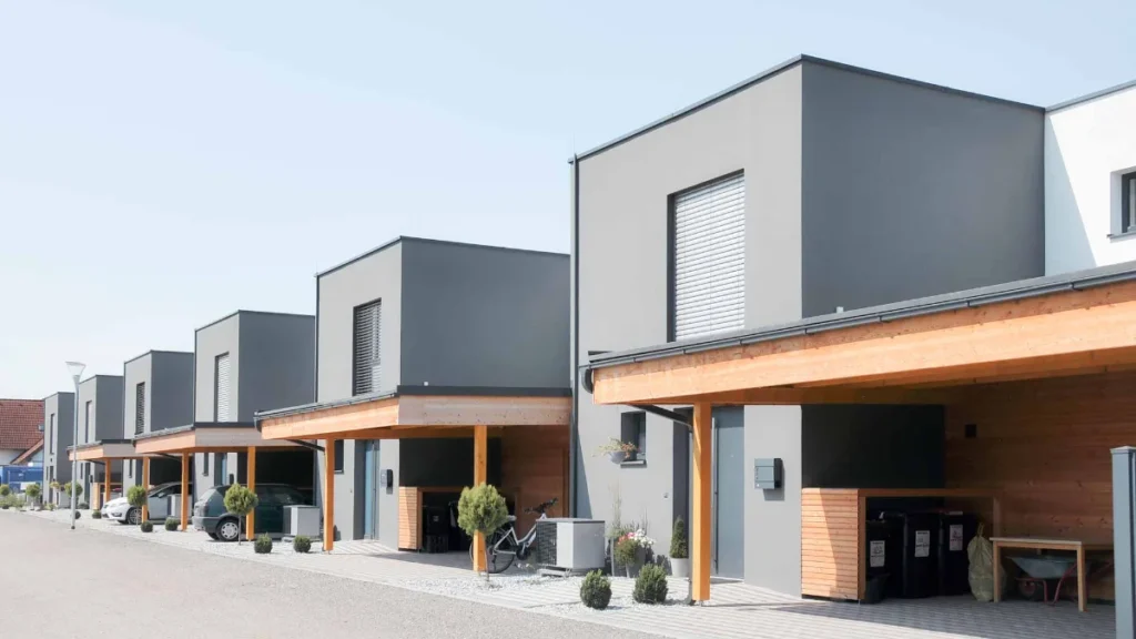 Eine Reihe moderner, kastenförmiger Häuser mit grauen Fassaden und hölzernen Akzenten, jedes mit einem überdachten Carport, kleinen Pflanzen und einer sauberen, gepflasterten Auffahrt an einem klaren Tag.