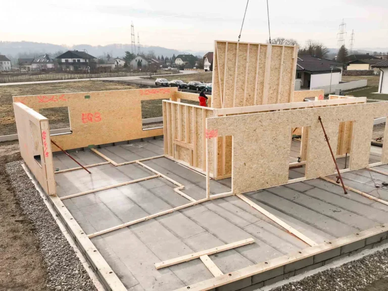 Auf einer Baustelle werden Holzrahmenwände auf einem Betonfundament errichtet, wobei Kräne und Arbeiter die Gebäudestruktur zusammensetzen. Im Hintergrund sind Häuser, Gras und Strommasten zu sehen.