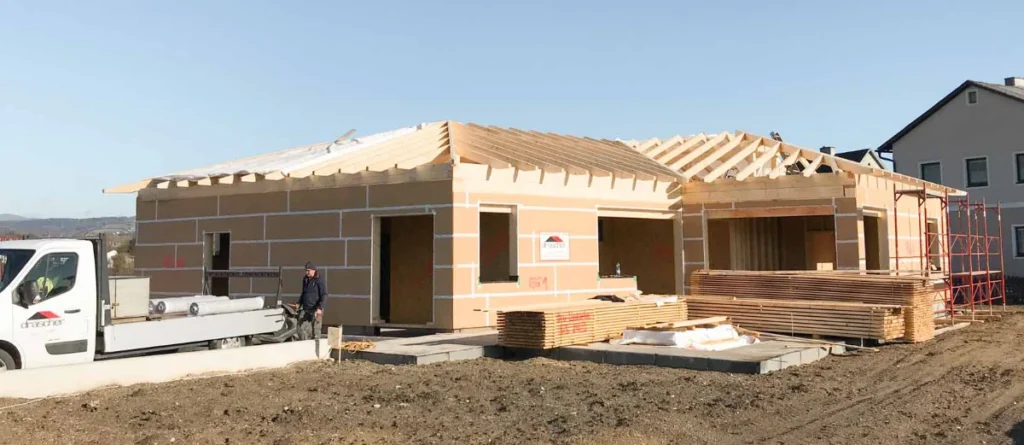 Ein einstöckiges, im Bau befindliches Haus mit freiliegendem Holzskelett und Dachstuhl, ein in der Nähe stehender Arbeiter, ein davor geparkter Lastwagen und Holzstapel auf dem Boden.