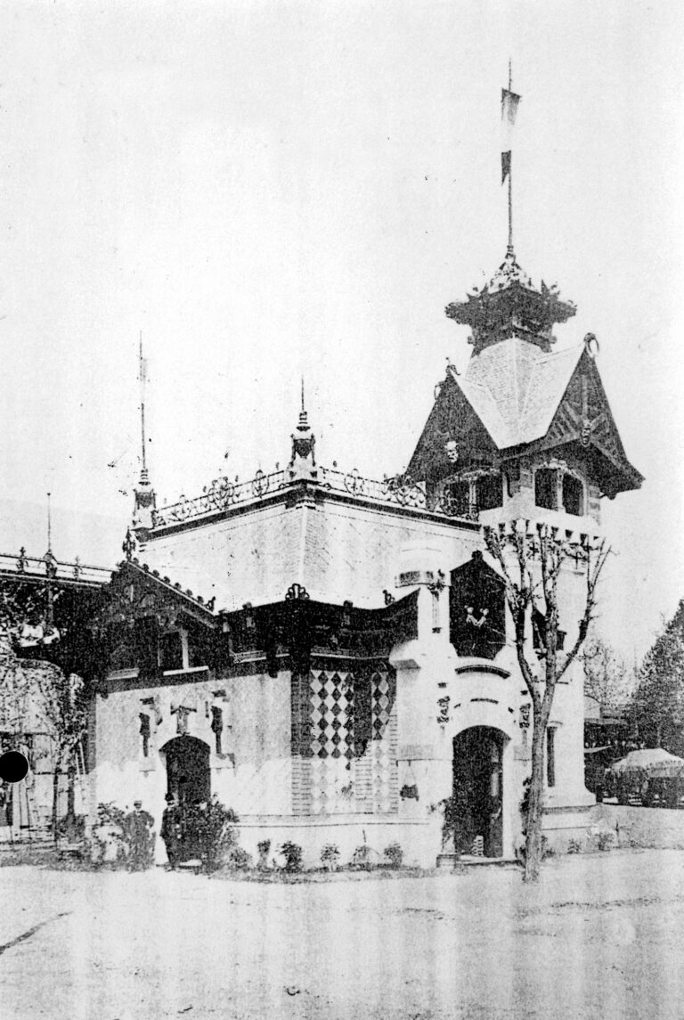 Schwarz-Weiß-Foto eines verschnörkelten Gebäudes mit dekorativem Dach, Türmchen und komplizierten architektonischen Details; Bäume und Sträucher säumen die Fassade, und auf dem Dach weht eine Flagge.