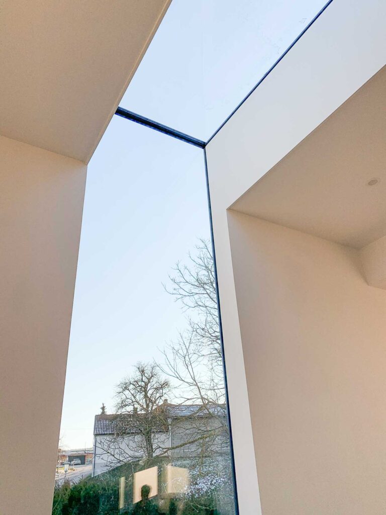 Blick aus dem Inneren eines modernen Gebäudes mit hohen weißen Wänden und einem großen Eckfenster, das den Blick auf einen klaren blauen Himmel und blattlose Bäume freigibt. Durch das Fenster ist ein Teil des Daches eines Hauses zu sehen.