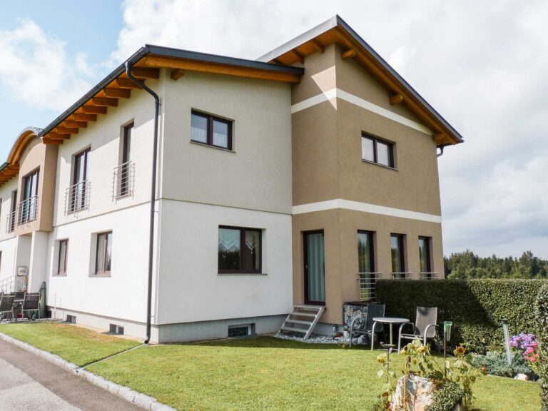 Ein modernes zweistöckiges Haus mit beigen und braunen Außenwänden, einem schrägen Dach und vielen Fenstern steht auf einem gepflegten grünen Rasen neben einer gepflasterten Straße unter einem teilweise bewölkten Himmel.