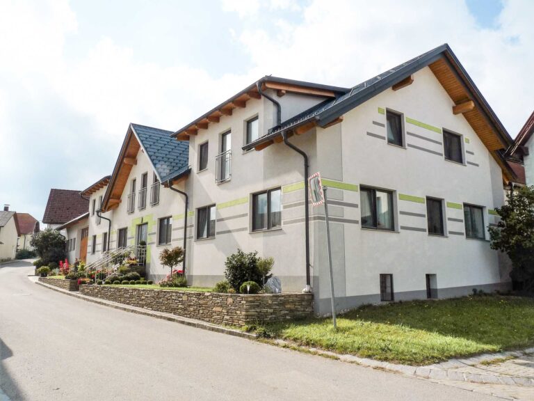 Ein modernes, helles Haus mit Schrägdach, grünen Akzenten und vielen Fenstern steht in einer ruhigen Wohnstraße mit einem kleinen Garten und einer Steinmauer davor.