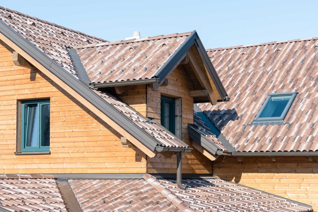 Holzhaus mit komplexem, geneigtem Ziegeldach mit Dachgauben und Oberlichtern, alles unter einem klaren blauen Himmel.
