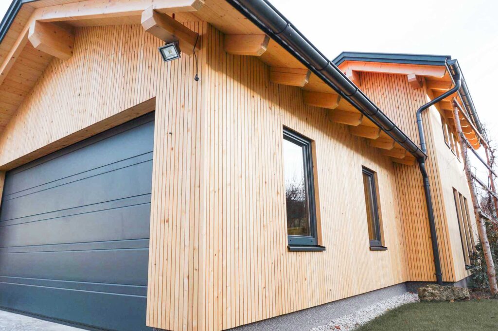 Modernes Holzhaus mit vertikaler heller Verkleidung, zwei schwarz gerahmten Fenstern, einem großen dunkelgrauen Garagentor und freiliegenden Dachbalken unter einem Schrägdach mit schwarzen Dachrinnen.