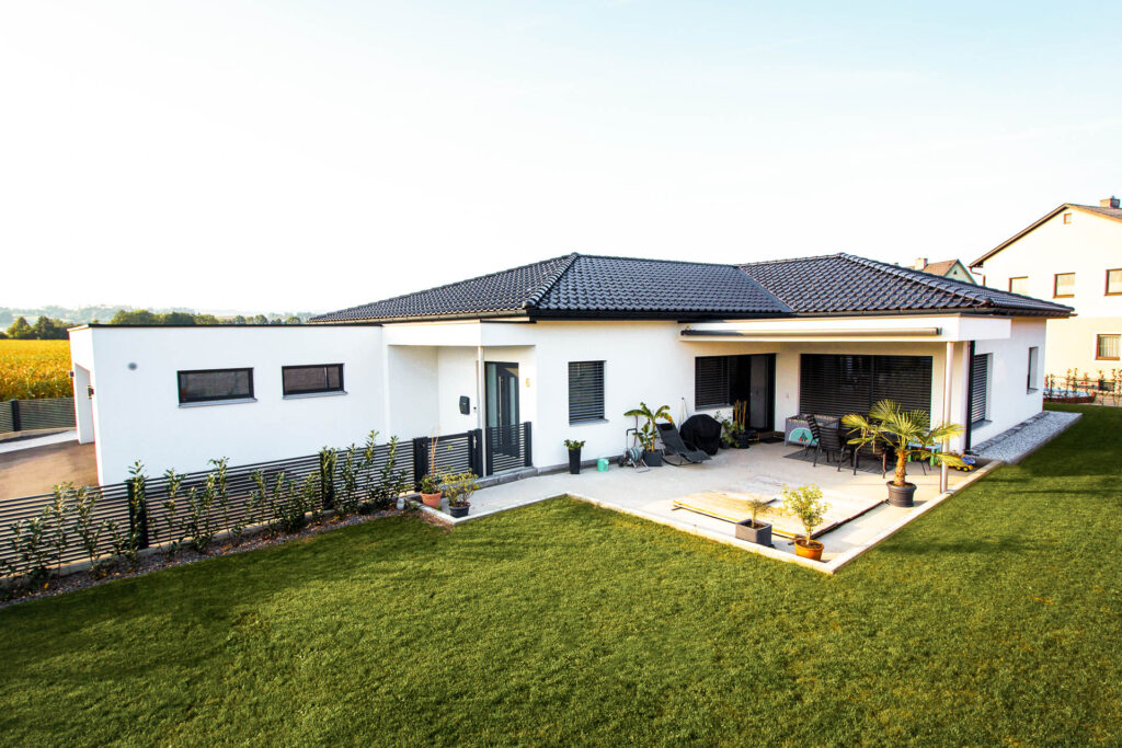 Modernes, einstöckiges, weißes Haus mit dunklem Ziegeldach, großen Fenstern, einer überdachten Terrasse mit Gartenmöbeln, Topfpflanzen und einem gepflegten grünen Rasen davor.