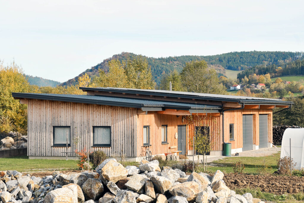 Ein modernes einstöckiges Holzhaus mit Schrägdach steht in einer ländlichen Gegend, umgeben von Felsen, grünem Gras und Bäumen, mit Hügeln und bewaldeter Landschaft im Hintergrund.
