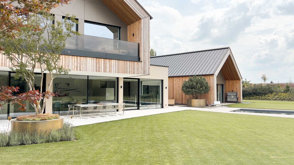Modernes Haus mit Holz- und Glasfassade, großen Fenstern und einer überdachten Terrasse mit Essplatz im Freien. Manicured Rasen, Baum in einem Pflanzer, und ein Schwimmbad sind in der geräumigen Hinterhof sichtbar.