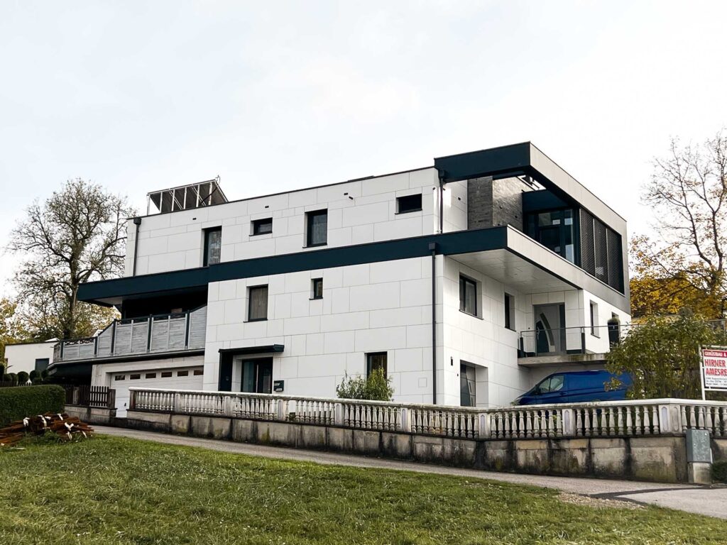 Ein modernes, weißes, mehrstöckiges Haus mit großen Fenstern, einem Balkon und einer Garage, umgeben von einem niedrigen Steinzaun und Grünflächen, unter einem bewölkten Himmel.