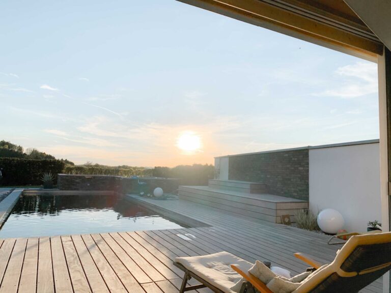Moderner Garten mit einer Holzterrasse, Liegestühlen und einem Pool bei Sonnenuntergang. Die Sonne steht tief am Himmel und wirft ein warmes Licht auf die friedliche Szene im Freien. Weiße kugelförmige Lichter und grüne Sträucher akzentuieren den Bereich.