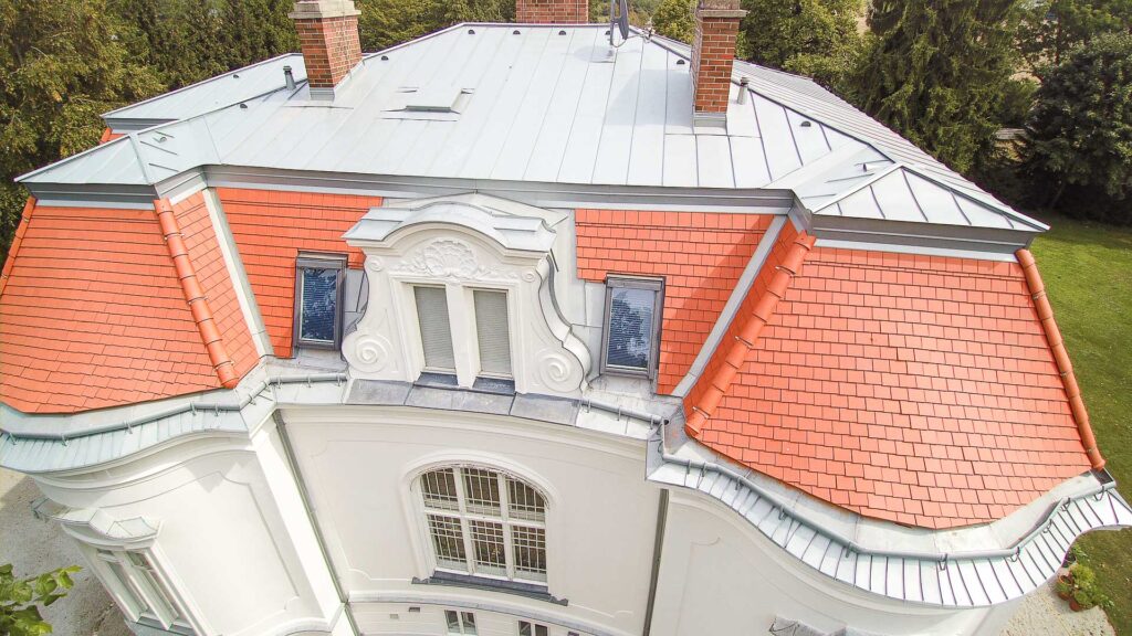 Luftaufnahme eines historischen Gebäudes mit rotgeziegeltem Mansarddach, weißer verzierter Fassade, zwei Schornsteinen und Dachgauben, umgeben von grünen Bäumen und Gras.