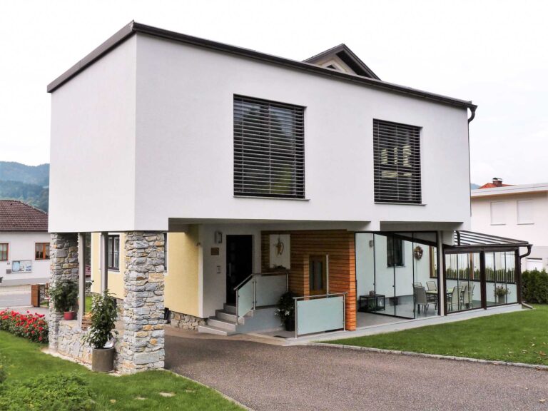 Modernes zweistöckiges Haus mit weißem, von Steinsäulen gestütztem Obergeschoss, großen Fenstern mit Jalousien, einem Untergeschoss mit Glaswänden und einem kleinen begrünten Hof mit Rasen und Blumen.