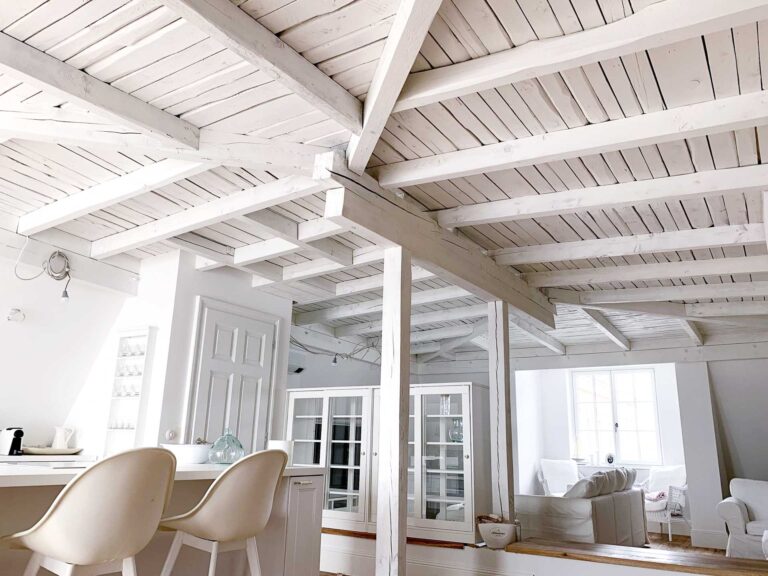 Ein heller, luftiger Raum mit weiß gestrichenen Holzbalken und einer Decke, weißen, modernen Stühlen an einem Tresen, Regalen, einer Glasvitrine und einem gemütlichen weißen Sofa unter einem großen Fenster.