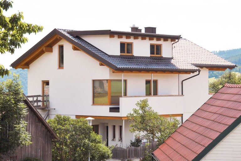 Ein modernes, weißes, zweistöckiges Haus mit dunklem Ziegeldach und Holzverkleidung, umgeben von Bäumen und benachbarten Häusern, vor dem Hintergrund grüner Hügel.