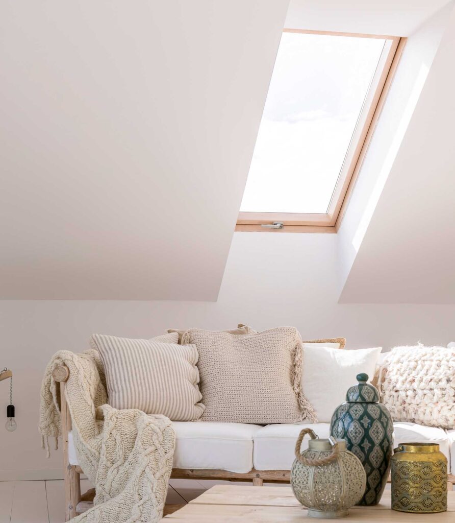 Ein gemütliches Zimmer im Dachgeschoss mit einem Oberlicht, einem weißen Sofa mit cremefarbenen Kissen und einer Strickdecke sowie dekorativen Vasen auf einem Holztisch davor. Sanftes natürliches Licht erfüllt den Raum.