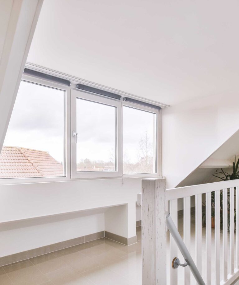 Helles, minimalistisches Innentreppenhaus mit weißen Wänden, Geländer, beigefarbenem Fliesenboden und großen Fenstern, die natürliches Licht hereinlassen; draußen sind ein Ziegeldach und der Himmel zu sehen, und in der Ecke steht eine Topfpflanze.