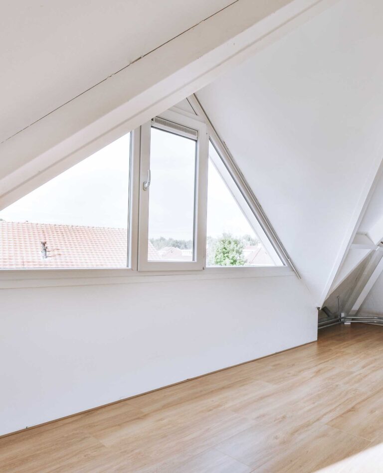 Ein helles Zimmer im Dachgeschoss mit schrägen weißen Wänden, einem großen dreieckigen Fenster und hellem Holzfußboden. Vor dem Fenster sind Dächer und Grünflächen zu sehen.