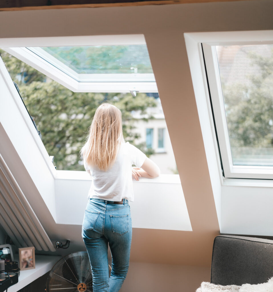 Eine Frau mit langen blonden Haaren, die ein weißes T-Shirt und blaue Jeans trägt, steht in einem modernen Dachzimmer und blickt durch ein offenes Fenster auf die Bäume und Gebäude draußen.