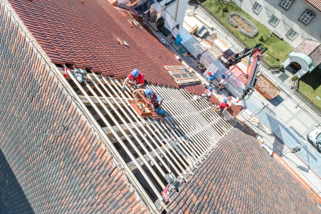 Arbeiter reparieren ein steiles Ziegeldach, wobei einige Ziegel entfernt werden, um Holzbalken freizulegen. Um das Gebäude herum sind Baumaschinen und -materialien zu sehen, und die Arbeiter tragen Sicherheitsausrüstung.