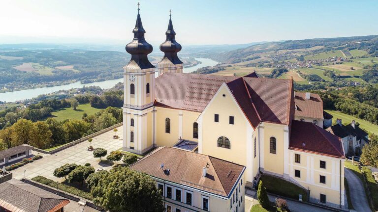 Eine große gelbe Barockkirche mit zwei Zwiebeltürmen steht auf einer Hügelkuppe mit Blick auf einen Fluss, grüne Felder und ferne Hügel unter einem klaren blauen Himmel.