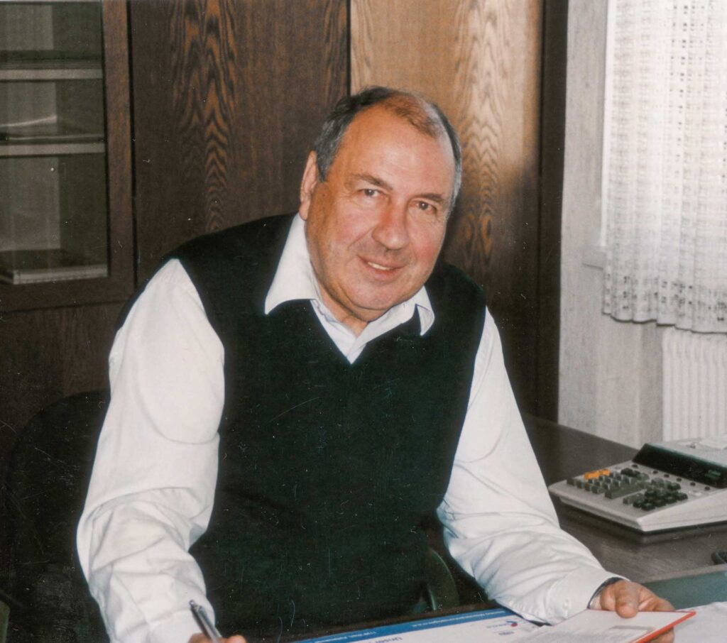 Ein älterer Mann mit kurzen grauen Haaren, einem weißen Hemd und einer dunklen Weste sitzt an einem Schreibtisch mit Papieren und einem Taschenrechner und lächelt in die Kamera. Im Hintergrund sind Holzschränke und ein Fenster mit Spitzenvorhängen zu sehen.