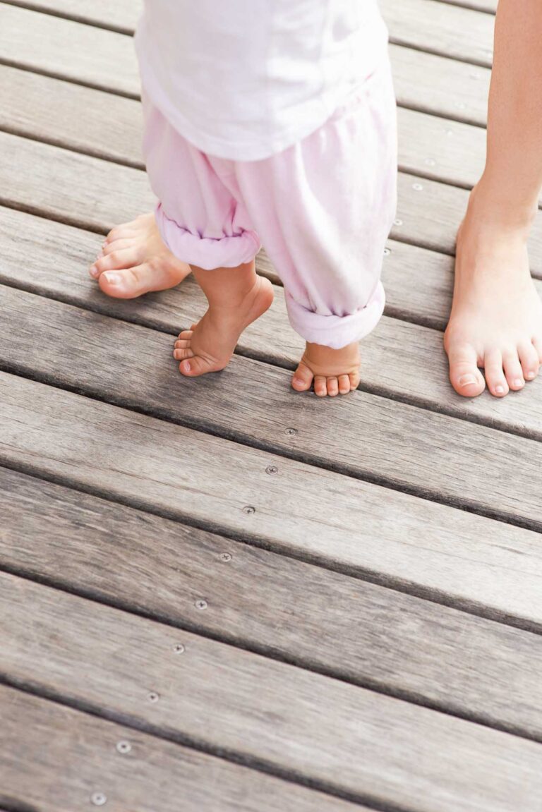 Ein Kleinkind in hellrosa Hosen steht barfuß auf einer Holzterrasse neben einem Erwachsenen, dessen Füße ebenfalls nackt und teilweise sichtbar sind.