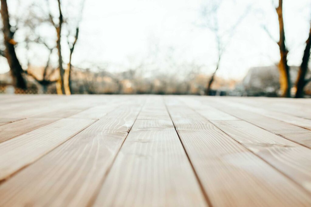 Nahaufnahme eines Holzdecks mit hellen Dielen, aufgenommen aus einem niedrigen Winkel. Im Hintergrund sind verschwommene Bäume und der Himmel zu sehen, was auf einen Außenbereich hindeutet.