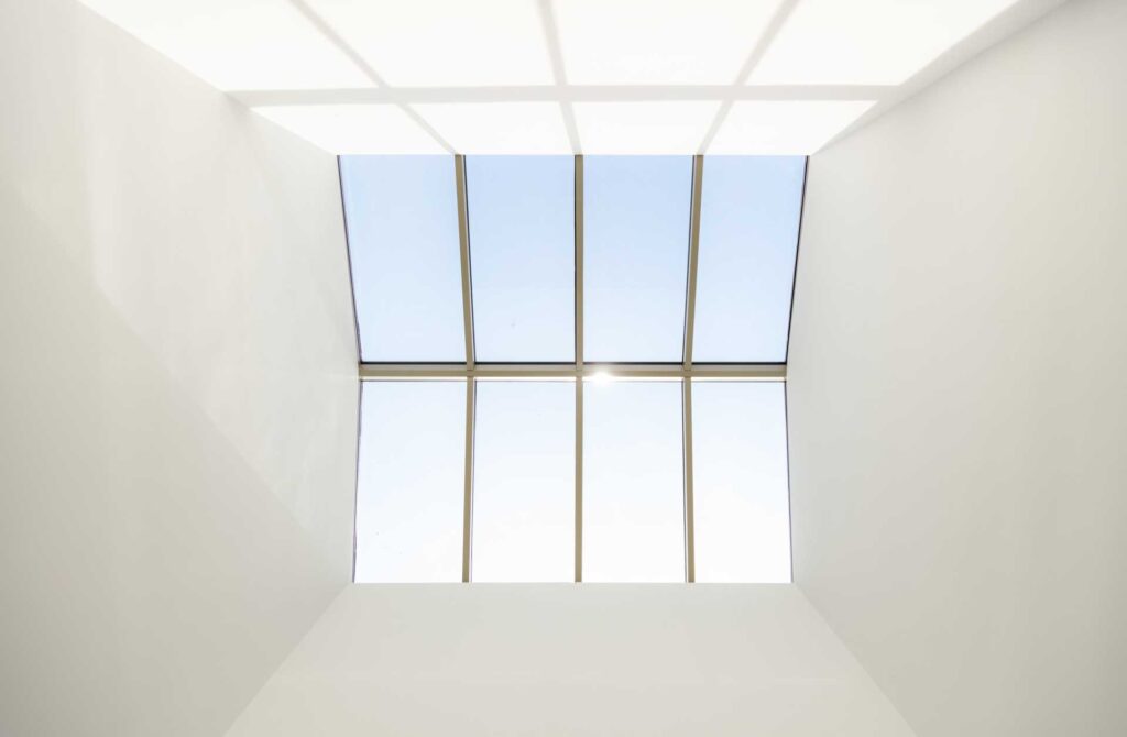 Ein großes rechteckiges Oberlicht mit sechs Glasscheiben und weißen Rahmen lässt Sonnenlicht in einen minimalistischen Raum mit weißen Wänden strömen. Der Himmel darüber ist klar und blau, und das Sonnenlicht wirft geometrische Schatten auf die Wände.