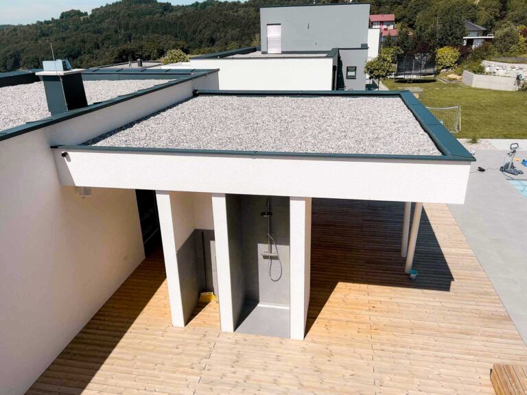 Eine moderne Außendusche mit minimalistischem Design ist unter einem Flachdach neben einer Holzterrasse gebaut; der Bereich grenzt an eine Rasenfläche und ein modernes Haus mit einem kiesbedeckten Dach.