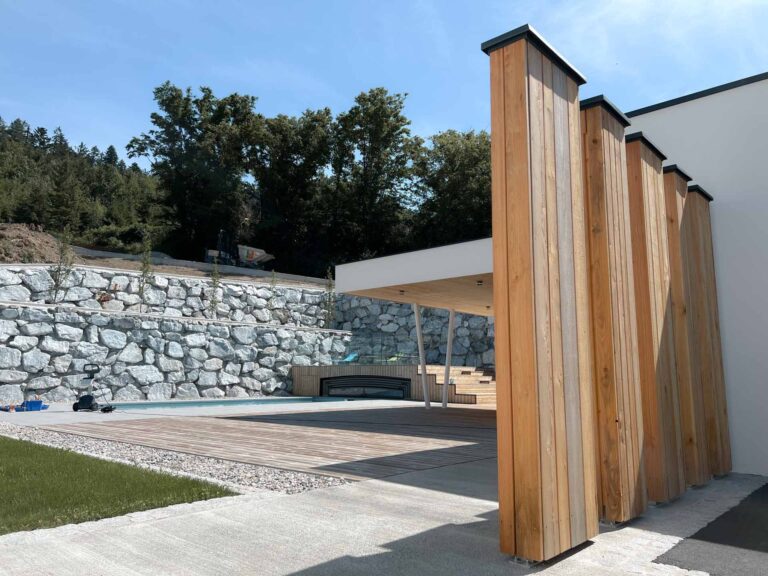 Ein moderner Außenbereich mit großen vertikalen Holzbalken, einer Holzterrasse und einer Steinmauer, umgeben von Bäumen und Grünpflanzen unter einem klaren blauen Himmel.