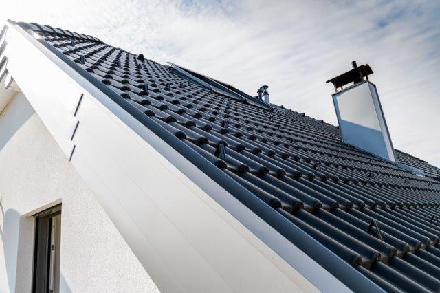 Nahaufnahme eines modernen Hauses mit einem Schrägdach mit dunkelgrauen Ziegeln, einem Dachfenster und einem Schornstein vor einem teilweise bewölkten Himmel.