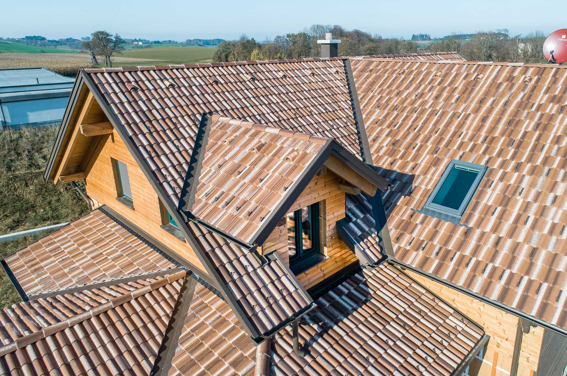 Luftaufnahme eines Hauses mit mehreren Spitzdächern, die mit braunen Ziegeln gedeckt sind, mit Oberlichtern und einer hölzernen Außenfassade, in einer ländlichen Landschaft mit Feldern und Bäumen im Hintergrund.