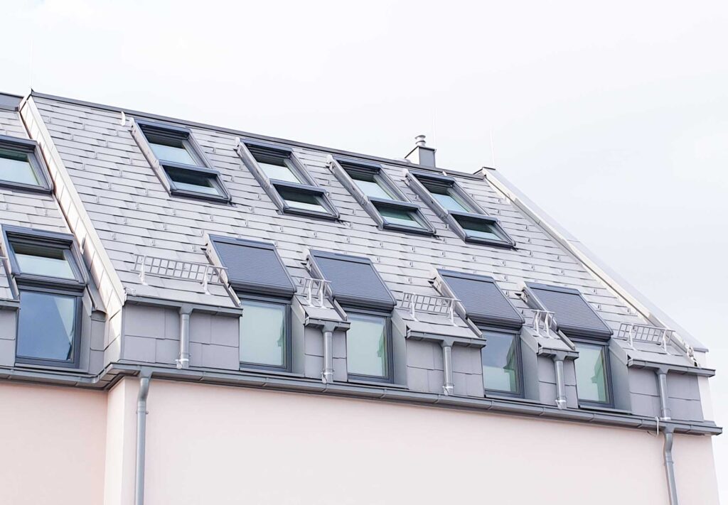 Ein modernes Gebäude mit einem schrägen grauen Dach mit mehreren Oberlichtfenstern und Balkonen auf dem Dach, von unten gesehen vor einem blassen Himmel.