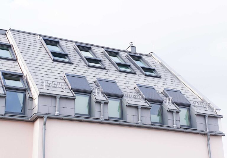 Ein modernes Gebäude mit einem schrägen grauen Dach mit mehreren Oberlichtfenstern und Balkonen auf dem Dach, von unten gesehen vor einem blassen Himmel.