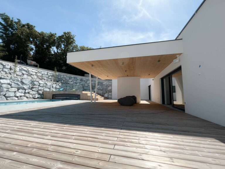 Ein modernes Haus mit einer großen Holzterrasse, die teilweise überdacht ist. Auf der Terrasse steht ein Sitzsack, und links ist ein Swimmingpool zu sehen. Im Hintergrund sind Steinmauern und Bäume unter einem blauen Himmel zu sehen.