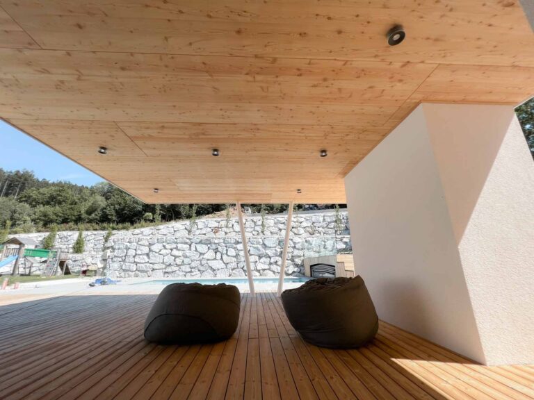 Zwei Sitzsäcke sitzen auf einer Holzterrasse unter einem modernen Überhang mit einer Holzdecke. Im Hintergrund sind eine Steinmauer und Grünflächen sowie ein Teil eines Swimmingpools zu sehen.