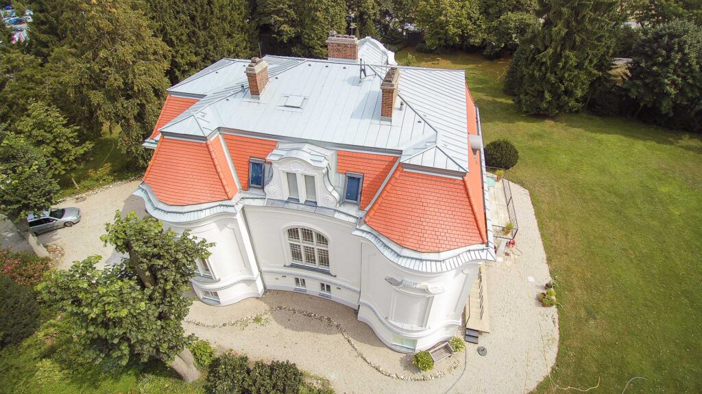 Luftaufnahme eines großen, eleganten weißen Hauses mit rotem Mansarddach, mehreren Schornsteinen und kunstvollen architektonischen Details, umgeben von grünem Rasen und Bäumen.