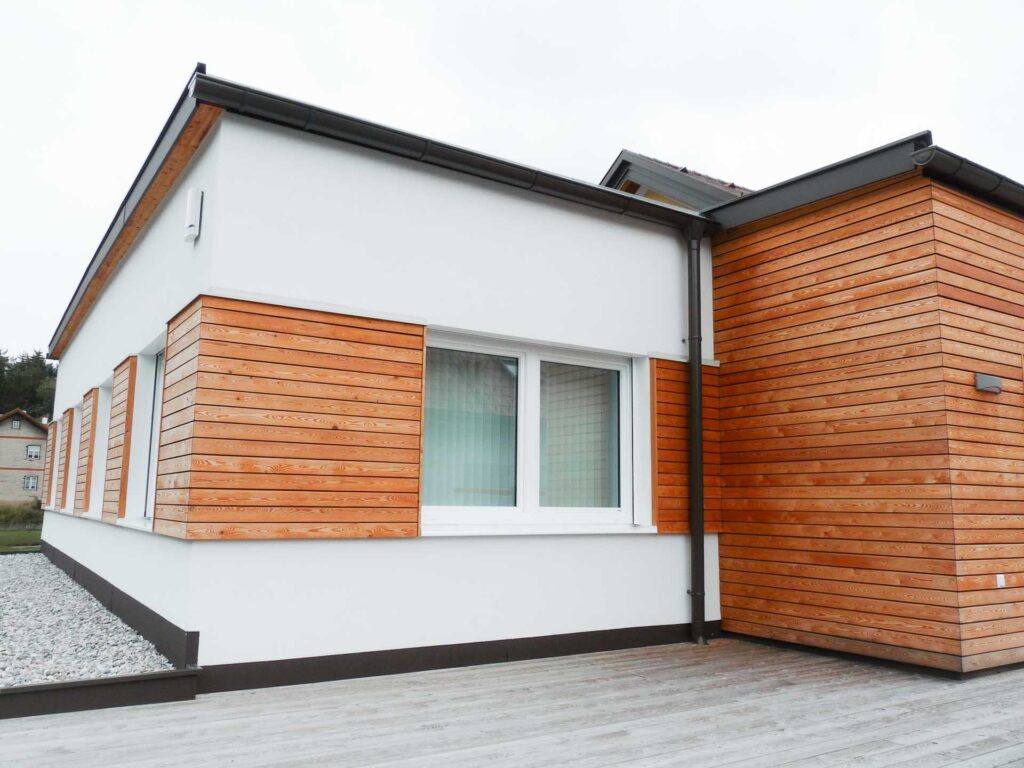 Modernes einstöckiges Haus mit weißen Wänden und horizontaler Holzverkleidung, mit einem großen Fenster und einem Kiesplatz neben einer Holzterrasse.