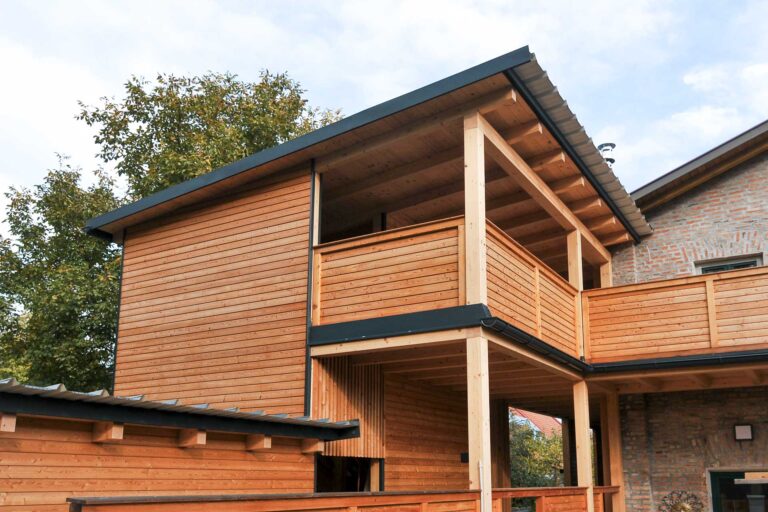 Ein modernes Haus mit einem oberen und einem unteren Stockwerk, mit Naturholzverkleidung, sichtbaren Balken und einem überdachten Balkon. Das Haus grenzt an ein Backsteingebäude und ist von Bäumen umgeben.