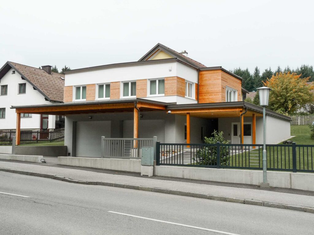 Ein modernes zweistöckiges Haus mit weißen Wänden, hölzernen Akzenten, großen Fenstern, einem angebauten Carport und einem gepflegten Rasen, gelegen an einer ruhigen Straße mit Nachbarhäusern und Bäumen im Hintergrund.