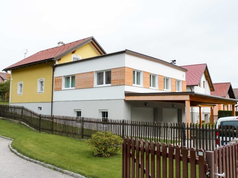 Ein modernes zweistöckiges Haus mit gelber und weißer Fassade, Ziegelsteinakzenten, einem schrägen roten Dach und einem Holzzaun davor. Das Haus hat einen gepflegten Rasen und liegt an einer leicht gebogenen Straße.