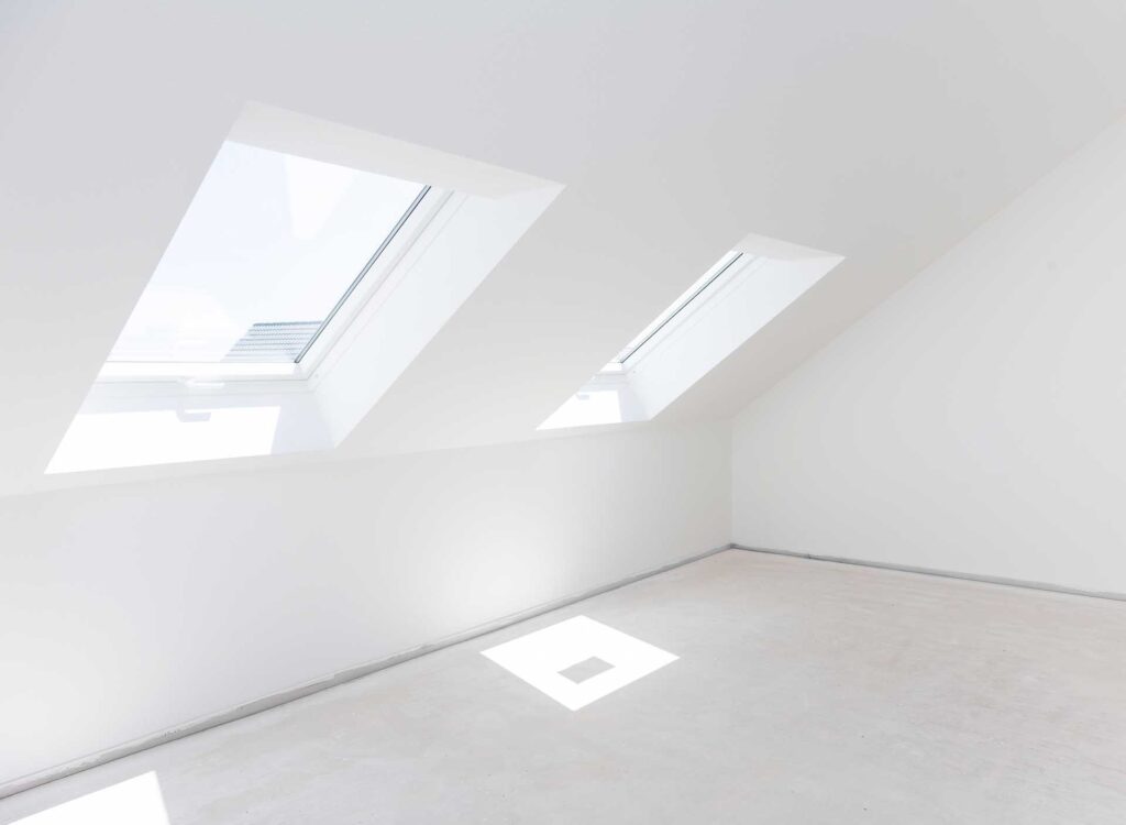 Helles, leeres Dachgeschosszimmer mit weißen Wänden und Decken, in dem drei große Dachfenster natürliches Licht hereinlassen und weiche Schatten auf den hellen Boden werfen.
