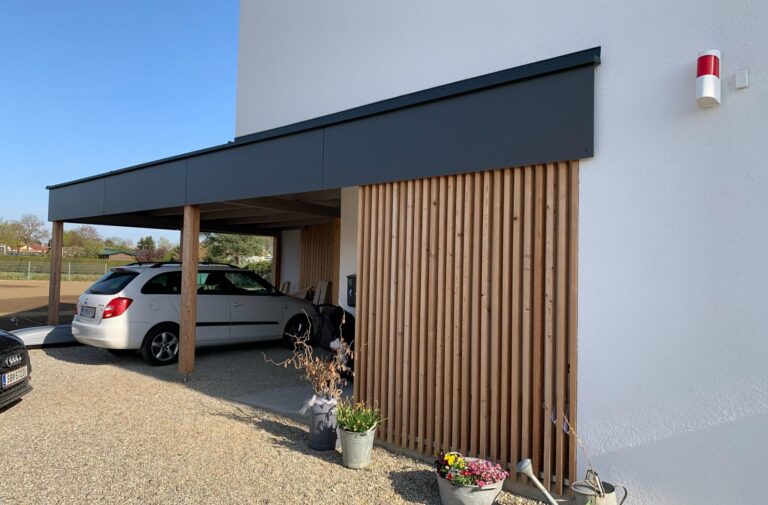 Ein moderner Carport, der an ein weißes Gebäude angebaut ist und ein silbernes Auto beherbergt. Die Struktur verfügt über vertikale Holzlatten, Kiesboden, Topfpflanzen und eine rot-weiße Außenleuchte an der Wand.