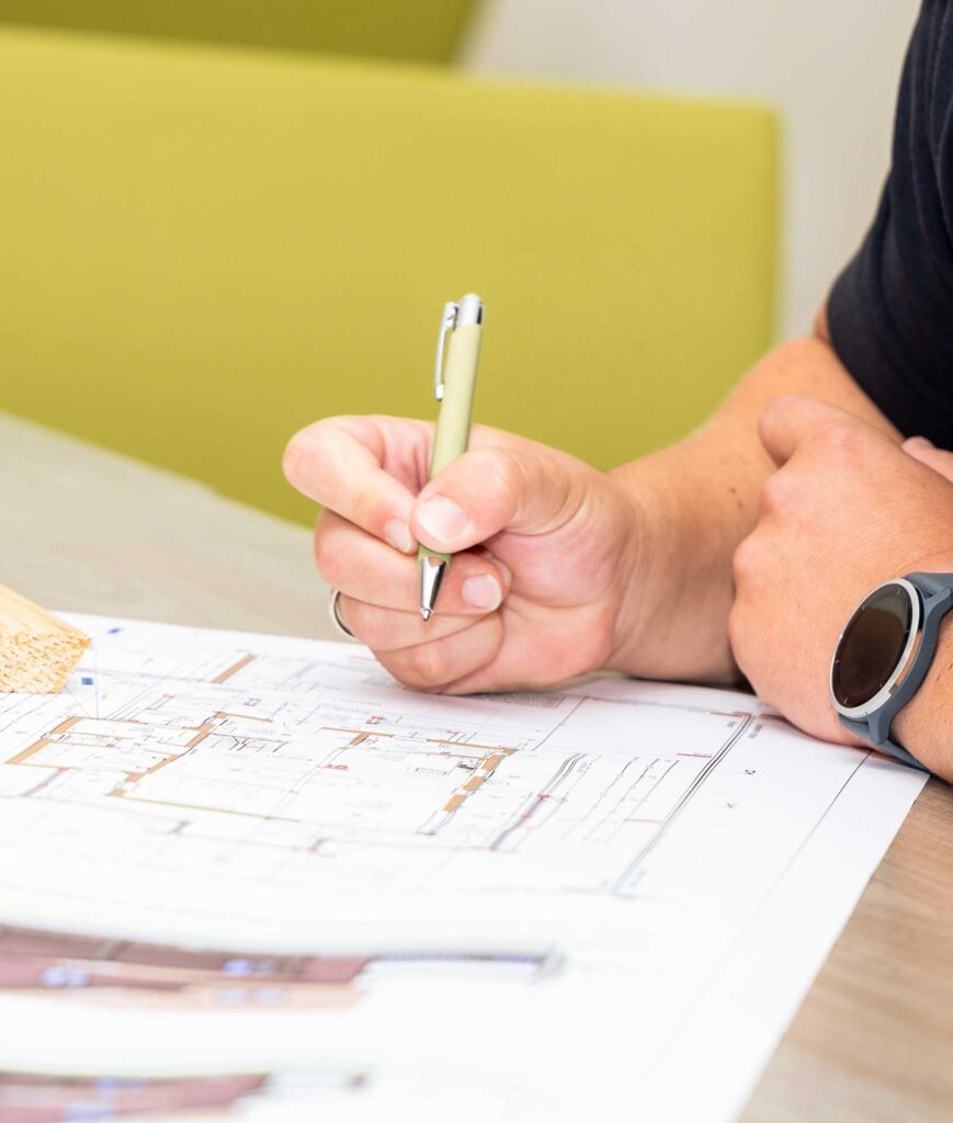 Eine Person, die eine Smartwatch trägt, skizziert oder schreibt mit einem Stift auf Architekturplänen, während sie an einem Tisch mit einem grünen Stuhl im Hintergrund sitzt.