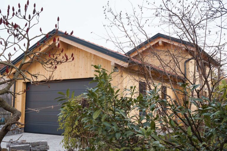 Ein modernes Holzhaus mit einem großen dunkelgrauen Garagentor, umgeben von grünen Bäumen und Sträuchern, von der Einfahrt aus gesehen.