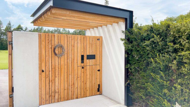 Modernes Holztor mit vertikalen Lamellen, einer kleinen Kranzverzierung, schwarzem Briefkasten und Gegensprechanlage, umrahmt von weißem Beton und Metall, neben hohen grünen Büschen unter einem teilweise bewölkten Himmel.